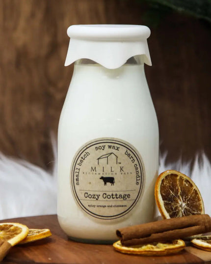 Milk bottle-shaped candle with 'Cozy Cottage' label on a wooden surface with dried oranges and cinnamon sticks.