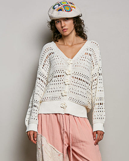 A woman modeling a cream-colored, open-knit crochet cardigan with decorative buttons, paired with a pink skirt and a colorful embroidered flat cap, against a plain light gray studio background.