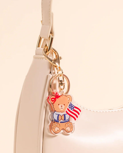 Teddy bear keychain with an American flag attached to a beige handbag.