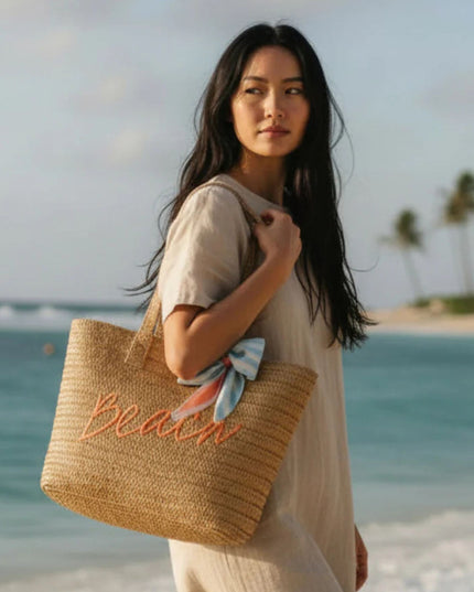 Woman holding a straw bag with 'Beach' embroidery on a beach.