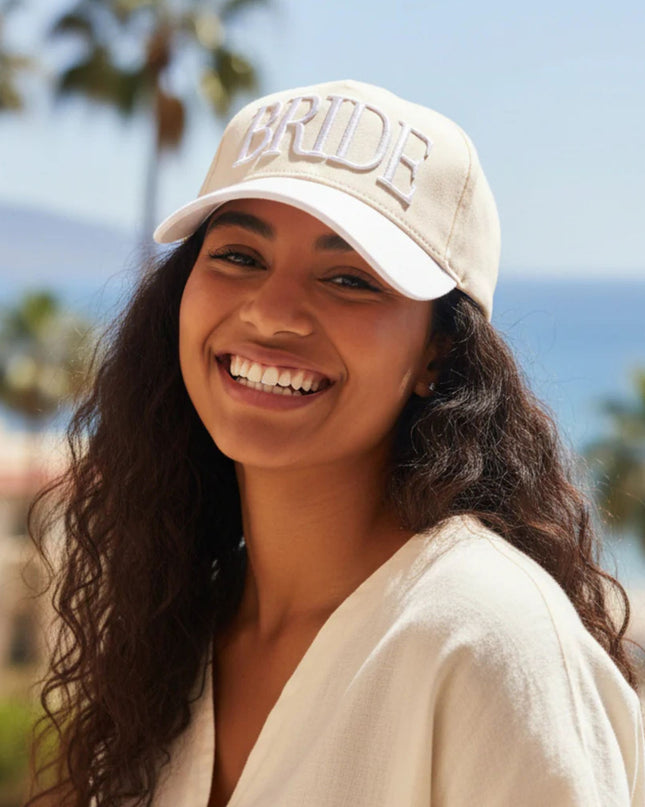Woman wearing a beige cap with 'BRIDE' text, smiling outdoors with palm trees and blue sky in the background.