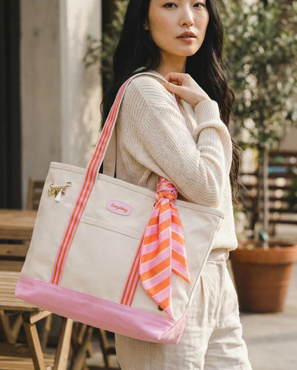 Woman holding a beige tote bag with pink accents and a striped scarf, standing outdoors.