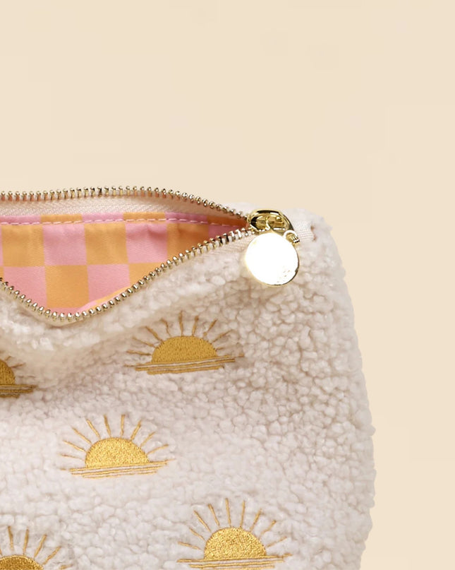 White pouch with gold sun designs and a pink and orange checkered zipper on a beige background