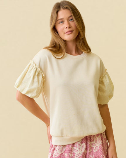 A woman modeling a cream-colored crewneck sweatshirt with puffed sleeves featuring ruffle trim, paired with pink patterned shorts, against a solid beige background.