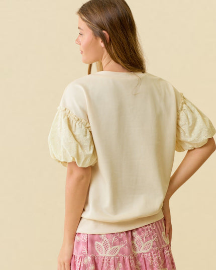 A woman shown from the back wearing a cream-colored short-sleeve top with textured, ruffled puff sleeves and pink patterned skirt. The background is a solid, warm beige studio backdrop.