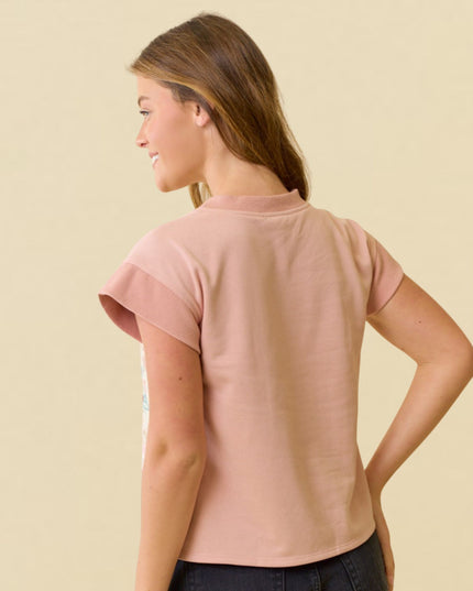 A woman shown from the back wearing a short-sleeve dusty pink t-shirt and dark jeans, standing against a solid light beige background.