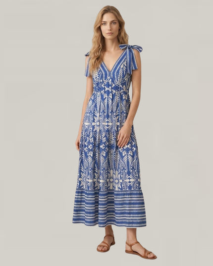 A woman modeling a blue and white patterned maxi dress with tie-shoulder straps and a V-neckline, paired with brown flat sandals, against a solid light gray background.