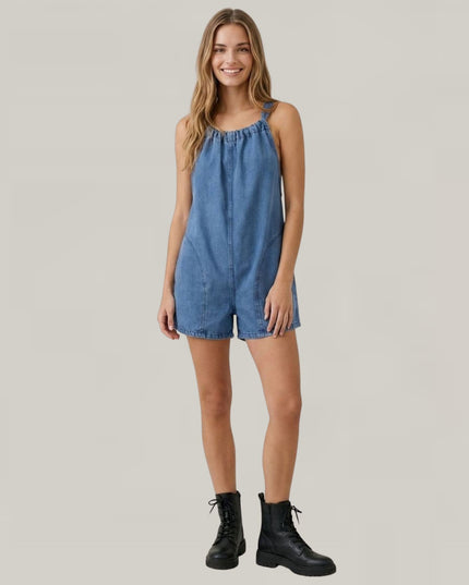 A woman modeling a sleeveless blue denim romper with a gathered neckline and short legs, paired with black lace-up ankle boots, against a solid light gray background.