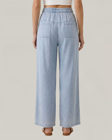 A model shown from the back wearing light blue wide-leg denim pants with an elastic waistband and two back patch pockets, set against a solid light gray background.