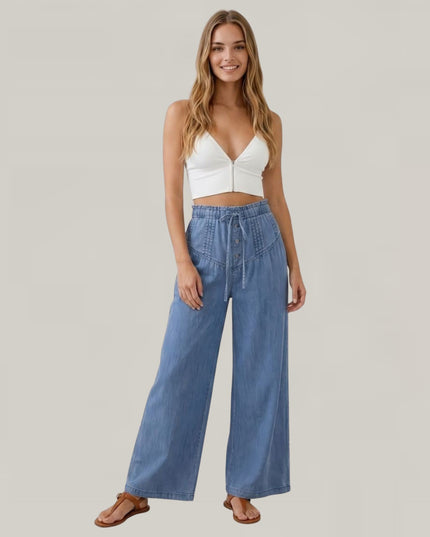 A woman modeling a white crop top and high-waisted, wide-leg blue denim jeans against a solid light gray background.