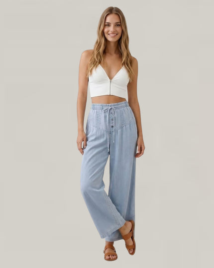 A woman modeling a white crop top and light blue wide-leg drawstring pants against a solid light gray background.