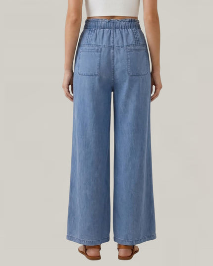 A model shown from the back wearing light blue wide-leg denim pants with an elastic waistband and back patch pockets, paired with a white crop top and brown sandals, against a solid light gray background.