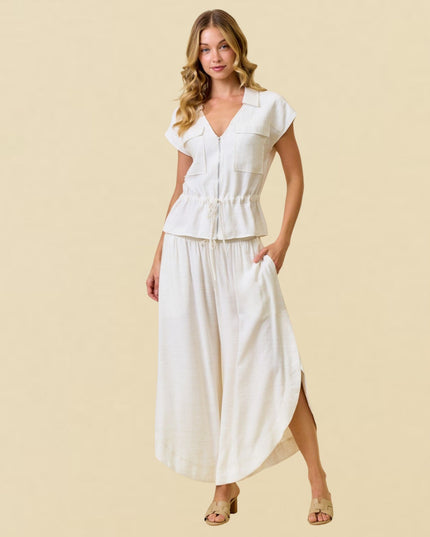 A woman modeling a white two-piece linen set consisting of a short-sleeve button-up top with a drawstring waist and matching wide-leg pants, set against a solid beige background.