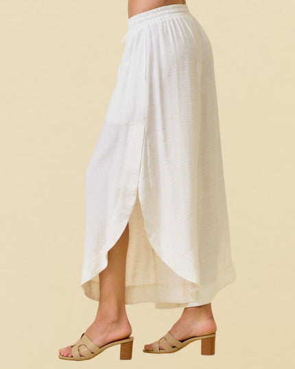 A model from the waist down wearing a white high-low midi skirt with a side slit and tan heeled mule sandals, set against a solid beige background.