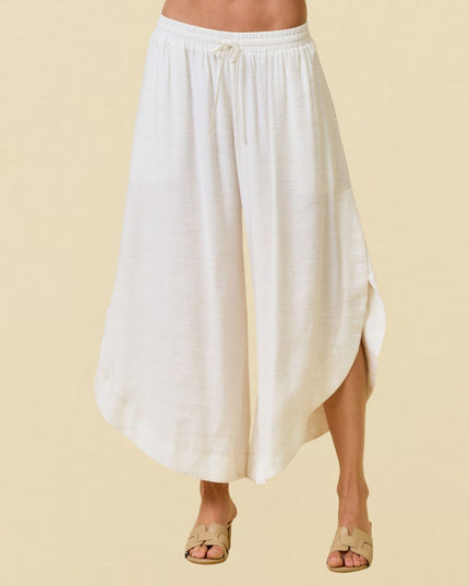 A person wearing a loose, high-waisted white maxi skirt with a drawstring waistband and side slit, paired with beige slide sandals, against a solid light beige background.