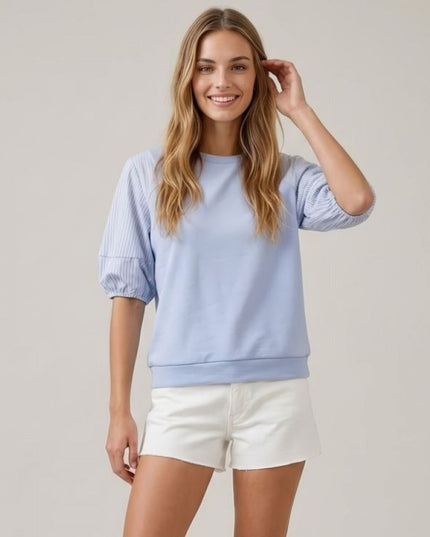 A woman modeling a light blue cropped sweatshirt with textured, vertically ribbed puff sleeves and white shorts against a plain light gray studio background.