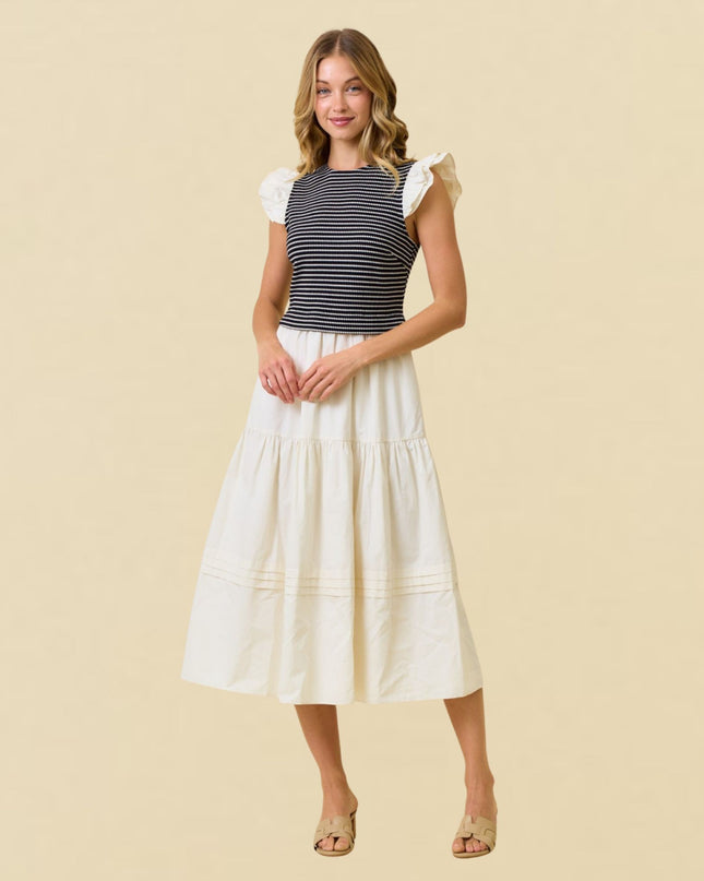 A woman modeling a two-piece outfit consisting of a navy and white striped short-sleeve top with ruffled sleeves and a matching cream-colored tiered midi skirt, set against a solid beige background.