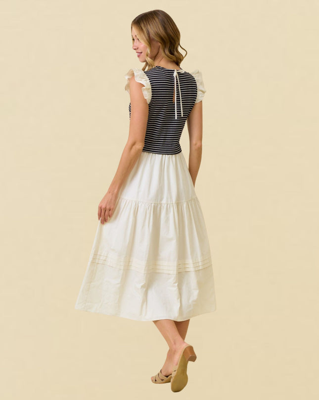 A woman modeling a sleeveless midi dress from the back, featuring a navy and white striped bodice with ruffled cap sleeves and a tiered off-white skirt, set against a solid beige background.