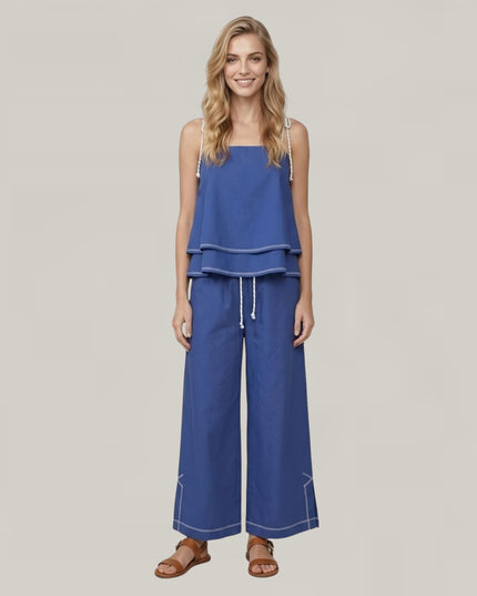 A woman modeling a blue sleeveless ruffled top and matching wide-leg pants set with white drawstrings and contrast stitching, photographed against a plain light gray studio background.
