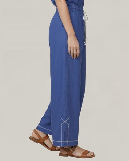 A person wearing wide-leg blue pants with white contrast stitching and a white drawstring waist, paired with brown flat sandals, shown against a plain light gray studio background.