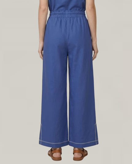A model shown from the back wearing a pair of wide-leg trousers in a medium blue color, paired with brown sandals, against a solid light gray background.