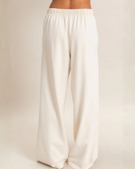 A person wearing loose, wide-leg pants in a warm off-white or cream color, shown from the waist down against a plain light beige studio background.