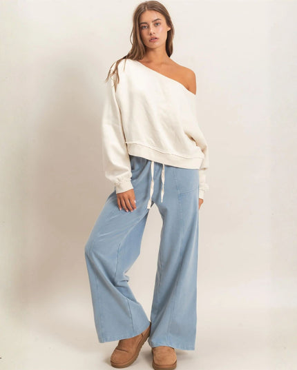 A woman modeling a cream off-the-shoulder long-sleeve top and light blue wide-leg drawstring pants, paired with tan slip-on shoes, against a plain off-white studio background.
