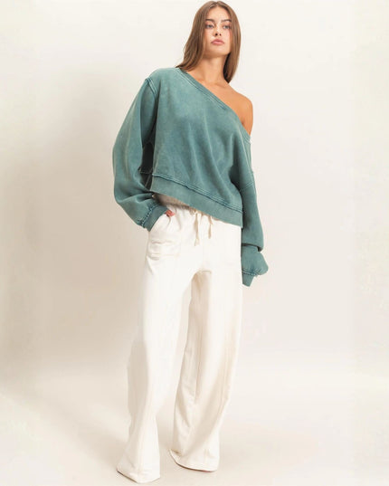 A woman modeling a teal green washed slouchy one-shoulder cropped sweatshirt paired with off-white wide-leg jeans against a plain off-white studio background.