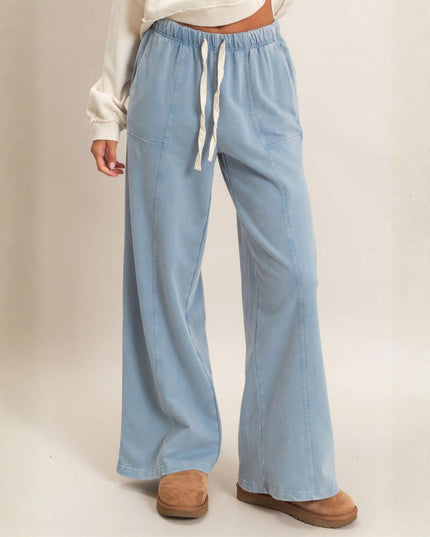 A person wearing light blue wide-leg drawstring pants with a white long-sleeve top and tan slip-on shoes, standing against a plain light beige studio background.