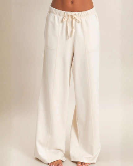 A person wearing off-white wide-leg lounge pants with an elastic drawstring waistband, shown from the torso down against a plain warm beige studio background.