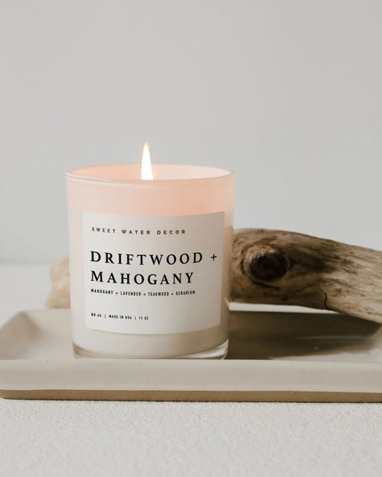 Candle labeled 'Driftwood + Mahogany' on a tray with driftwood on a light background