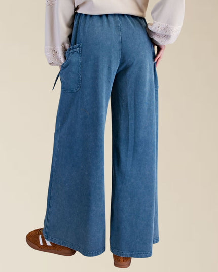 A person shown from the back wearing wide-leg blue denim pants with side pockets, a light beige blouse, and brown slip-on shoes against a plain beige studio background.