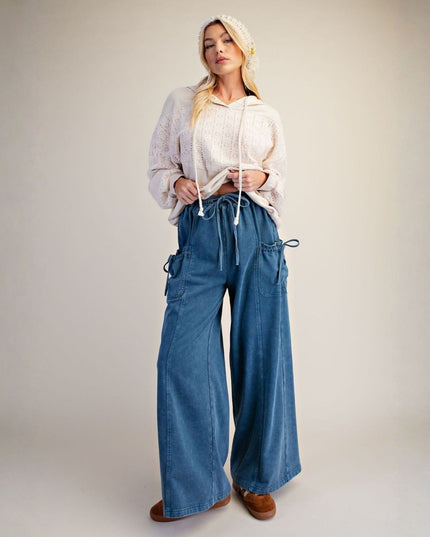 A woman stands against a plain light beige studio background wearing a cream knit cropped hoodie, wide-leg blue denim cargo pants, a cream beanie, and brown shoes.