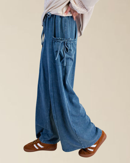 A person wearing wide-leg blue denim pants with drawstring waist and side pockets, paired with brown suede sneakers, shown from the waist down against a plain beige background.