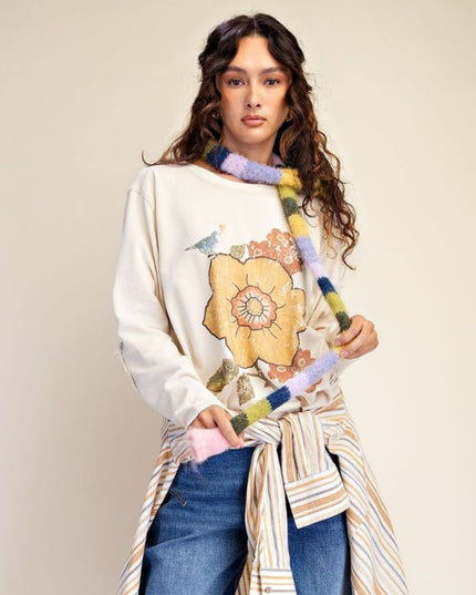 A woman modeling a cream-colored sweatshirt with a large yellow and orange floral graphic on the front, styled with a striped shirt tied around her waist and blue jeans. She is holding a colorful fuzzy knit scarf against a plain beige studio background.