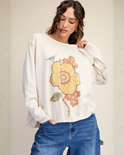 A woman wearing a cream-colored long-sleeve top with a large yellow and orange floral graphic on the front, paired with blue jeans, standing against a neutral beige background.