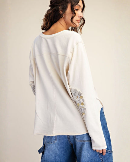 A woman shown from the back wearing a cream-colored long-sleeve top with decorative floral embroidery on the sleeves and blue jeans, set against a plain beige studio background.