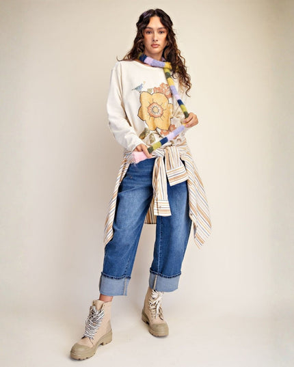 A model stands against a plain beige studio background wearing a cream sweatshirt with a large yellow floral graphic, blue jeans, and beige combat boots. She is styled with a striped shirt tied around her waist and a colorful patterned scarf.