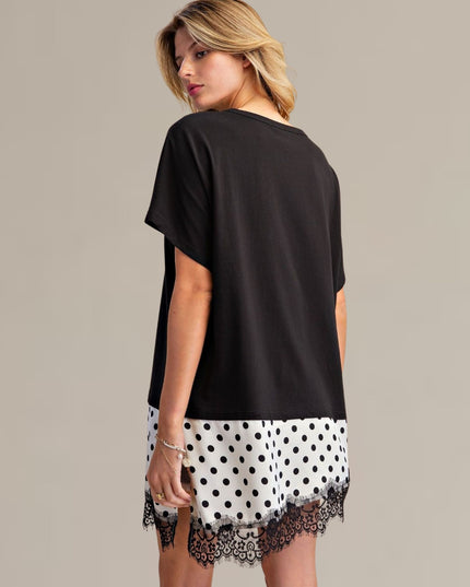 A woman modeling a short-sleeve black tunic dress with a white and black polka dot hem and black lace trim, shown from a three-quarters back view against a solid beige studio background.