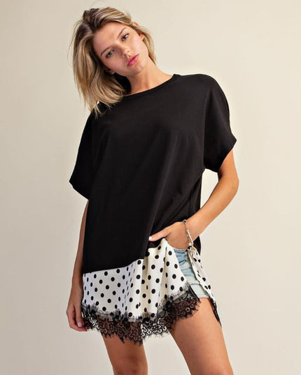 A woman modeling a loose black short-sleeve top paired with white shorts featuring black polka dots and black lace trim, against a plain beige studio background.