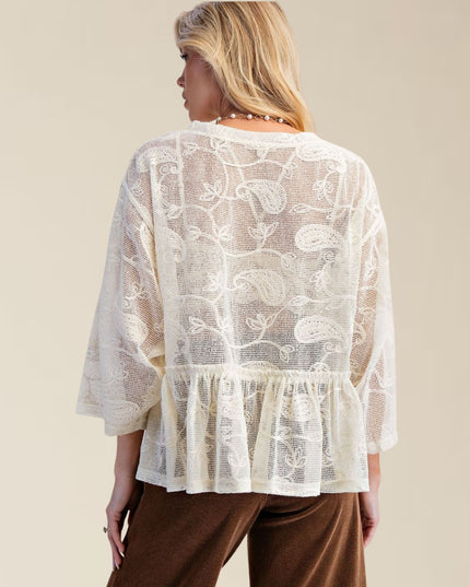 A woman shown from the back wearing a cream-colored lace top with paisley embroidery and brown pants, set against a solid beige studio background.