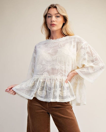 A woman modeling a white sheer lace blouse with wide bell sleeves and a ruffled peplum hem, paired with brown pants, against a plain beige studio background.