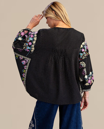 A woman shown from the back wearing a black long-sleeve blouse with white polka dots and colorful floral embroidery on the sleeves, paired with blue jeans, against a solid beige background.