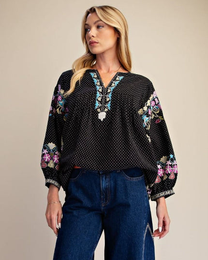 A woman modeling a black long-sleeve blouse with white polka dots, colorful floral embroidery on the sleeves, and a V-neckline with white beadwork, paired with blue jeans against a plain beige background.