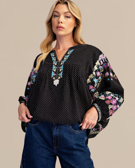 A woman modeling a black long-sleeve blouse with white polka dots, colorful floral embroidery on the sleeves and neckline, and blue jeans against a solid beige background.