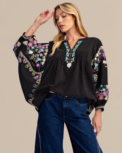 A woman modeling a black long-sleeve blouse with white polka dots, colorful floral embroidery on the sleeves and neckline, and a split V-neckline, paired with dark blue jeans against a solid beige background.