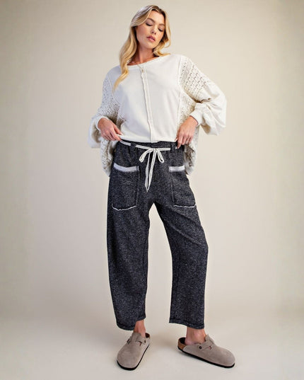 A woman modeling a pair of dark gray textured wide-leg pants with white contrast stitching and a white drawstring waist, styled with a white knit top and taupe slip-on shoes against a neutral beige studio background.