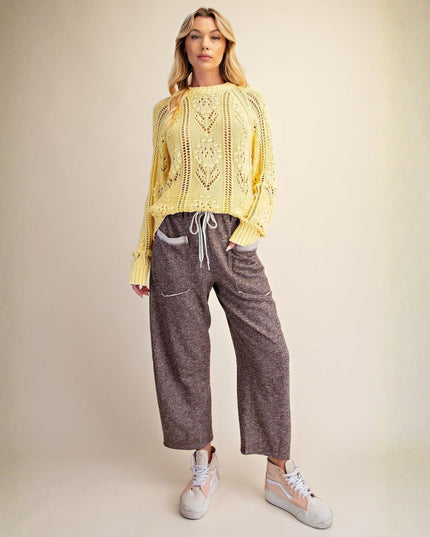 A woman modeling a pale yellow open-knit sweater and heathered brown wide-leg cropped pants with white sneakers, standing against a plain beige studio backdrop.