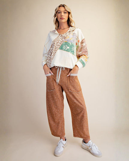 A woman stands against a plain beige studio background wearing a white cropped hoodie with multicolor abstract patches, brown wide-leg cropped pants, and white sneakers.