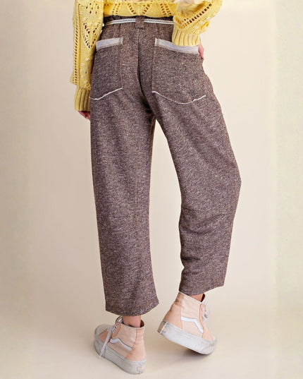 A person shown from the back wearing loose, cropped brown tweed trousers with white contrast stitching and back pockets, paired with a yellow knit top and light pink sneakers against a plain beige studio background.